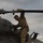 A soldier in MultiCam/OCP camouflage uniform is performing maintenance or inspection on top of a military helicopter. The aircraft has 'Kentucky' written on its fuselage, indicating it belongs to the Kentucky National Guard. The soldier is gripping a main rotor blade while positioned on top of the aircraft under an overcast sky.