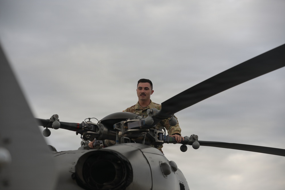 A military service member in camouflage uniform is positioned atop a helicopter, appearing to inspect or perform maintenance on the rotor hub assembly. The individual is standing or crouching near the main rotor head of what appears to be a military helicopter. The image is taken from a low angle, with an overcast sky in the background.