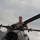 A military service member in camouflage uniform is positioned atop a helicopter, appearing to inspect or perform maintenance on the rotor hub assembly. The individual is standing or crouching near the main rotor head of what appears to be a military helicopter. The image is taken from a low angle, with an overcast sky in the background.