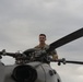 A military service member in camouflage uniform is positioned atop a helicopter, appearing to inspect or perform maintenance on the rotor hub assembly. The individual is standing or crouching near the main rotor head of what appears to be a military helicopter. The image is taken from a low angle, with an overcast sky in the background.