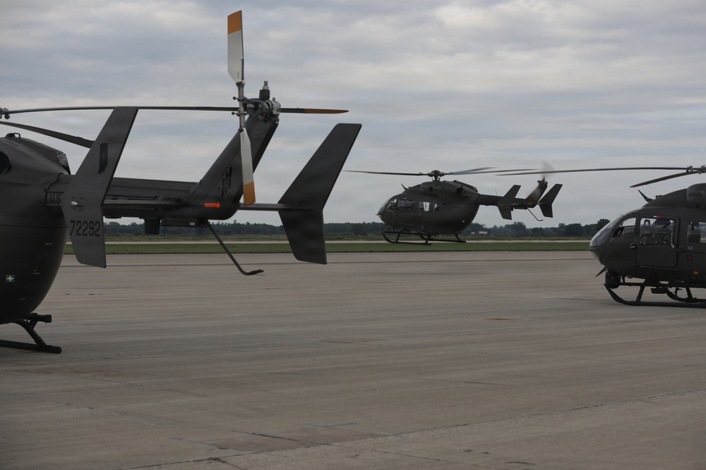 The image shows multiple military helicopters on a tarmac or airfield under an overcast sky. At least three UH-72 Lakota helicopters are visible, with one appearing to be in the process of landing or hovering in the background. The helicopters are painted in standard U.S. Army olive drab/dark gray livery.