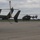 The image shows multiple military helicopters on a tarmac or airfield under an overcast sky. At least three UH-72 Lakota helicopters are visible, with one appearing to be in the process of landing or hovering in the background. The helicopters are painted in standard U.S. Army olive drab/dark gray livery.