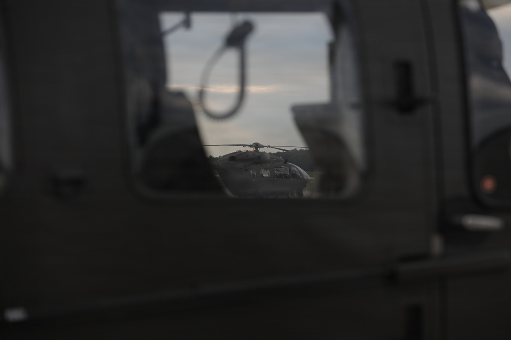 The image is taken from inside or alongside a military vehicle, looking through an opening or window toward a helicopter on the ground. The scene appears to be at dusk or dawn, with low ambient light creating a dark, moody atmosphere. A helicopter, likely military, is visible in the background framed by vehicle components.