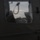 The image is taken from inside or alongside a military vehicle, looking through an opening or window toward a helicopter on the ground. The scene appears to be at dusk or dawn, with low ambient light creating a dark, moody atmosphere. A helicopter, likely military, is visible in the background framed by vehicle components.