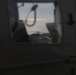 The image is taken from inside or alongside a military vehicle, looking through an opening or window toward a helicopter on the ground. The scene appears to be at dusk or dawn, with low ambient light creating a dark, moody atmosphere. A helicopter, likely military, is visible in the background framed by vehicle components.