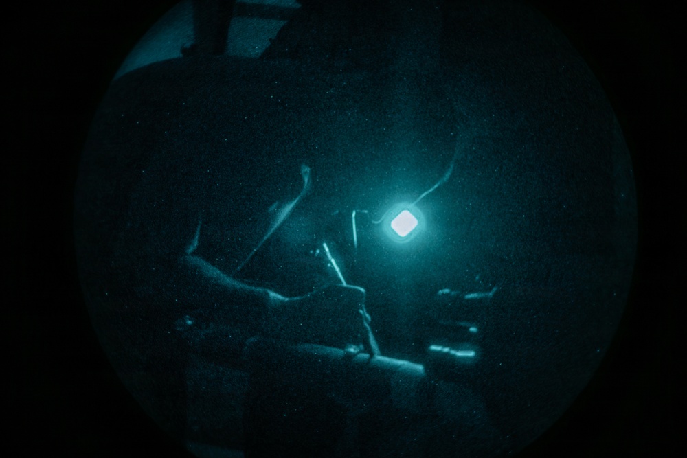 A night vision or low-light image showing a person working with what appears to be a long tool or weapon in a dark environment. A bright light source, possibly a headlamp or tactical light, is visible near the subject's head area. The circular vignette framing is characteristic of night vision optics or a similar optical device.