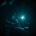 A night vision or low-light image showing a person working with what appears to be a long tool or weapon in a dark environment. A bright light source, possibly a headlamp or tactical light, is visible near the subject's head area. The circular vignette framing is characteristic of night vision optics or a similar optical device.