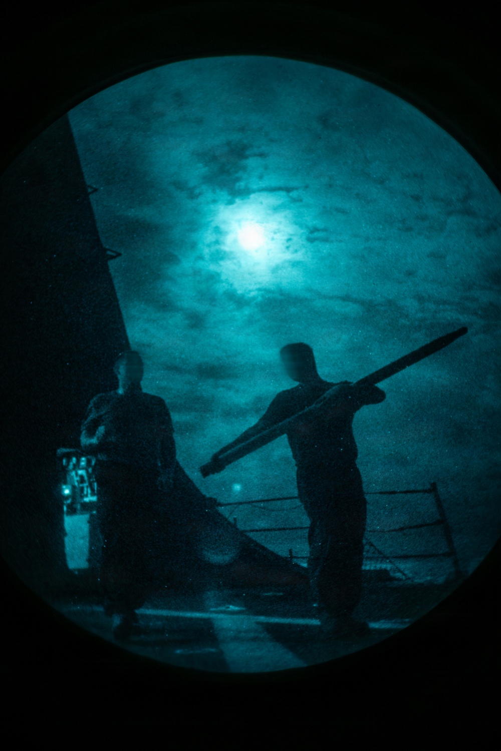 A night-vision or image-intensified photograph showing two silhouetted figures on what appears to be the deck of a naval vessel. One figure is carrying a long cylindrical object, possibly a weapon component or equipment. The scene is illuminated by a bright light source (likely the moon) through cloud cover, rendered in characteristic green-teal night-vision tones.