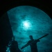 A night-vision or image-intensified photograph showing two silhouetted figures on what appears to be the deck of a naval vessel. One figure is carrying a long cylindrical object, possibly a weapon component or equipment. The scene is illuminated by a bright light source (likely the moon) through cloud cover, rendered in characteristic green-teal night-vision tones.