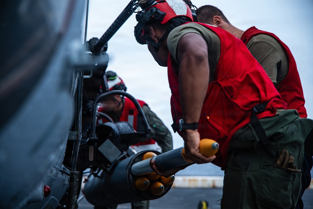 Military ordnance personnel wearing red vests and flight deck helmets are loading or inspecting rocket munitions on an aircraft, likely aboard a naval vessel. At least two crew members are visible working on what appears to be a rocket pod mounted to a military helicopter or aircraft. The setting appears to be a flight deck environment based on the surface texture and equipment visible.