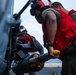 Military ordnance personnel wearing red vests and flight deck helmets are loading or inspecting rocket munitions on an aircraft, likely aboard a naval vessel. At least two crew members are visible working on what appears to be a rocket pod mounted to a military helicopter or aircraft. The setting appears to be a flight deck environment based on the surface texture and equipment visible.