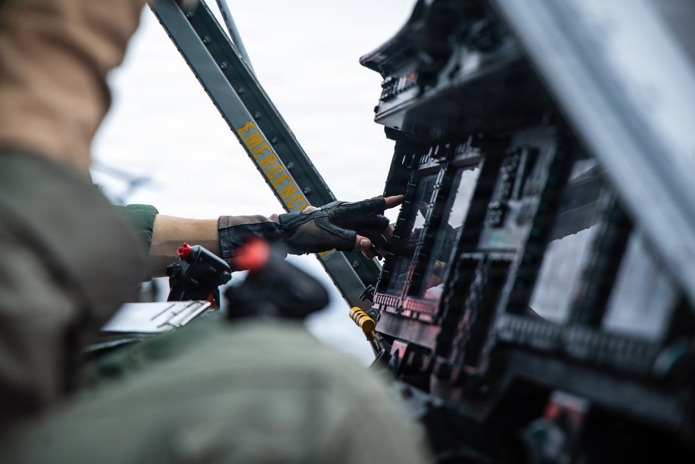 A military aviator wearing a flight suit and fingerless leather gloves is operating controls inside an aircraft cockpit. The image captures a close-up view of the pilot's hand interacting with instrument panels and switches. A yellow 'EMERGENCY' label is prominently visible on a structural beam within the cockpit.