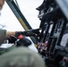 A military aviator wearing a flight suit and fingerless leather gloves is operating controls inside an aircraft cockpit. The image captures a close-up view of the pilot's hand interacting with instrument panels and switches. A yellow 'EMERGENCY' label is prominently visible on a structural beam within the cockpit.
