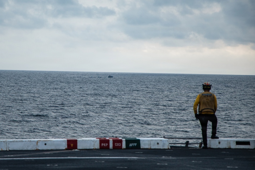 A crew member in a yellow jersey and helmet stands at the edge of an aircraft carrier flight deck, looking out toward the open ocean. A small vessel is visible on the horizon in the distance. The flight deck barrier displays fire suppression agent labels including PKP, CO2, and AFFF.