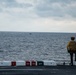 A crew member in a yellow jersey and helmet stands at the edge of an aircraft carrier flight deck, looking out toward the open ocean. A small vessel is visible on the horizon in the distance. The flight deck barrier displays fire suppression agent labels including PKP, CO2, and AFFF.
