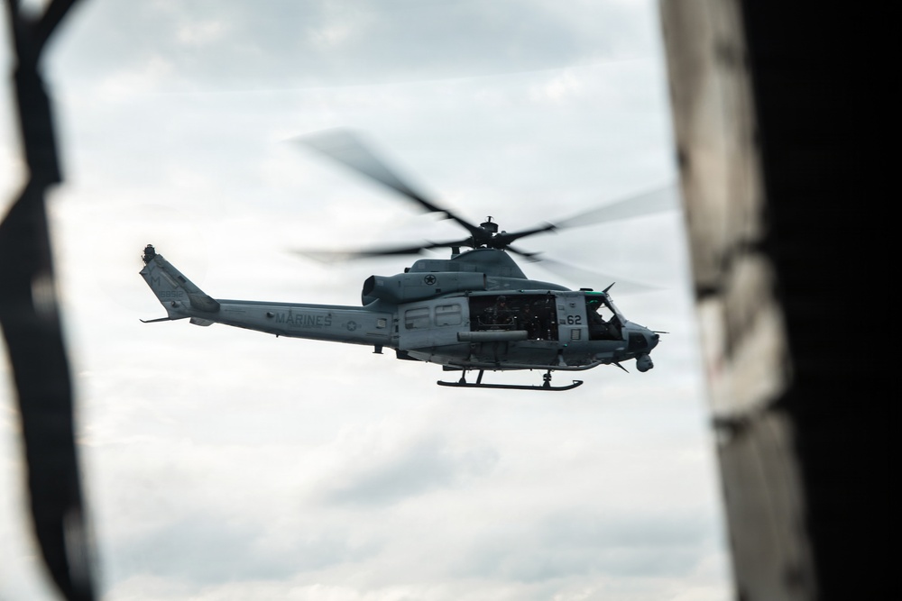 A U.S. Marines military helicopter, marked with the number '62' and 'MARINES' on its fuselage, is captured in flight against an overcast sky. The image is taken from inside another aircraft or structure, with dark framing elements visible on the left and right edges. The helicopter appears to be a Bell UH-1Y Venom or similar variant used by the United States Marine Corps.
