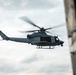 A U.S. Marines military helicopter, marked with the number '62' and 'MARINES' on its fuselage, is captured in flight against an overcast sky. The image is taken from inside another aircraft or structure, with dark framing elements visible on the left and right edges. The helicopter appears to be a Bell UH-1Y Venom or similar variant used by the United States Marine Corps.