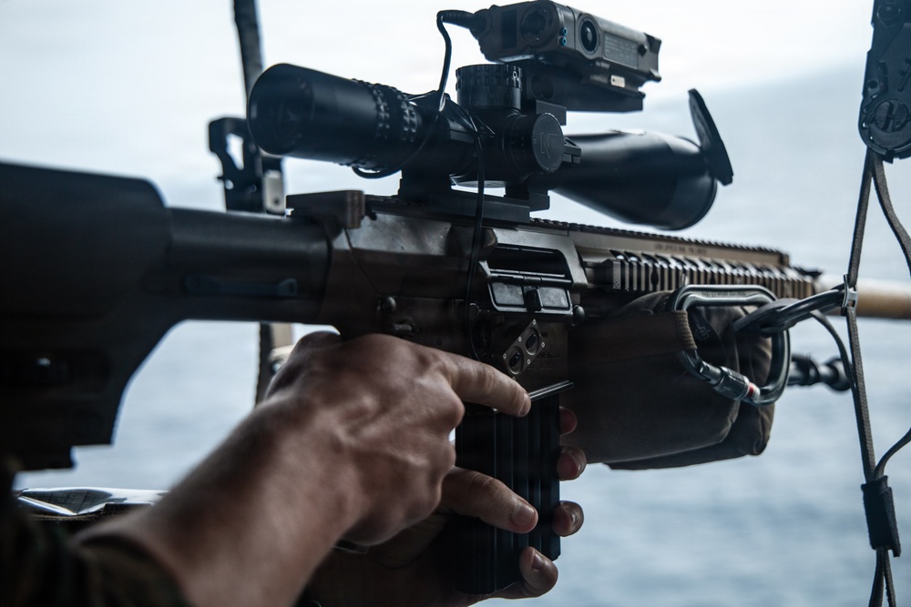 A close-up photograph of a military operator handling a modern assault rifle with multiple optics attachments. The weapon is equipped with a magnified rifle scope, a laser/illuminator device, and Picatinny rail accessories. The setting appears to be aboard a naval vessel or watercraft, indicated by the visible ocean in the background.