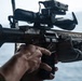 A close-up photograph of a military operator handling a modern assault rifle with multiple optics attachments. The weapon is equipped with a magnified rifle scope, a laser/illuminator device, and Picatinny rail accessories. The setting appears to be aboard a naval vessel or watercraft, indicated by the visible ocean in the background.