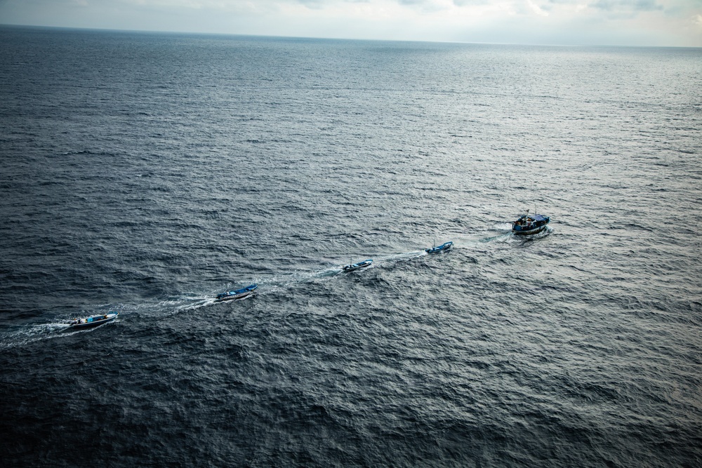 An aerial photograph shows a larger vessel towing or leading a chain of three smaller boats across open ocean. The boats are arranged in a line, each creating visible wake patterns in the dark blue water. The image is taken from a significant altitude, likely from an aircraft or helicopter.
