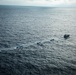 An aerial photograph shows a larger vessel towing or leading a chain of three smaller boats across open ocean. The boats are arranged in a line, each creating visible wake patterns in the dark blue water. The image is taken from a significant altitude, likely from an aircraft or helicopter.