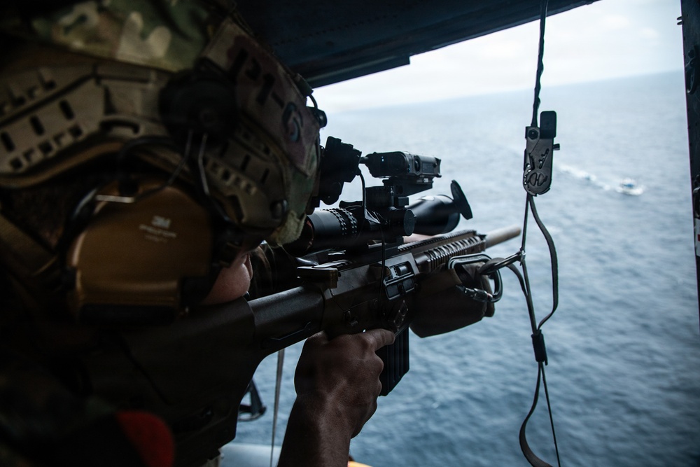 A military operator is positioned at the open door of an aircraft, aiming a rifle equipped with optical sighting systems toward the ocean below. The individual is wearing full tactical gear including a multicam helmet with mounted devices and hearing protection. A small vessel or wake is visible on the water surface in the background.