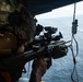 A military operator is positioned at the open door of an aircraft, aiming a rifle equipped with optical sighting systems toward the ocean below. The individual is wearing full tactical gear including a multicam helmet with mounted devices and hearing protection. A small vessel or wake is visible on the water surface in the background.