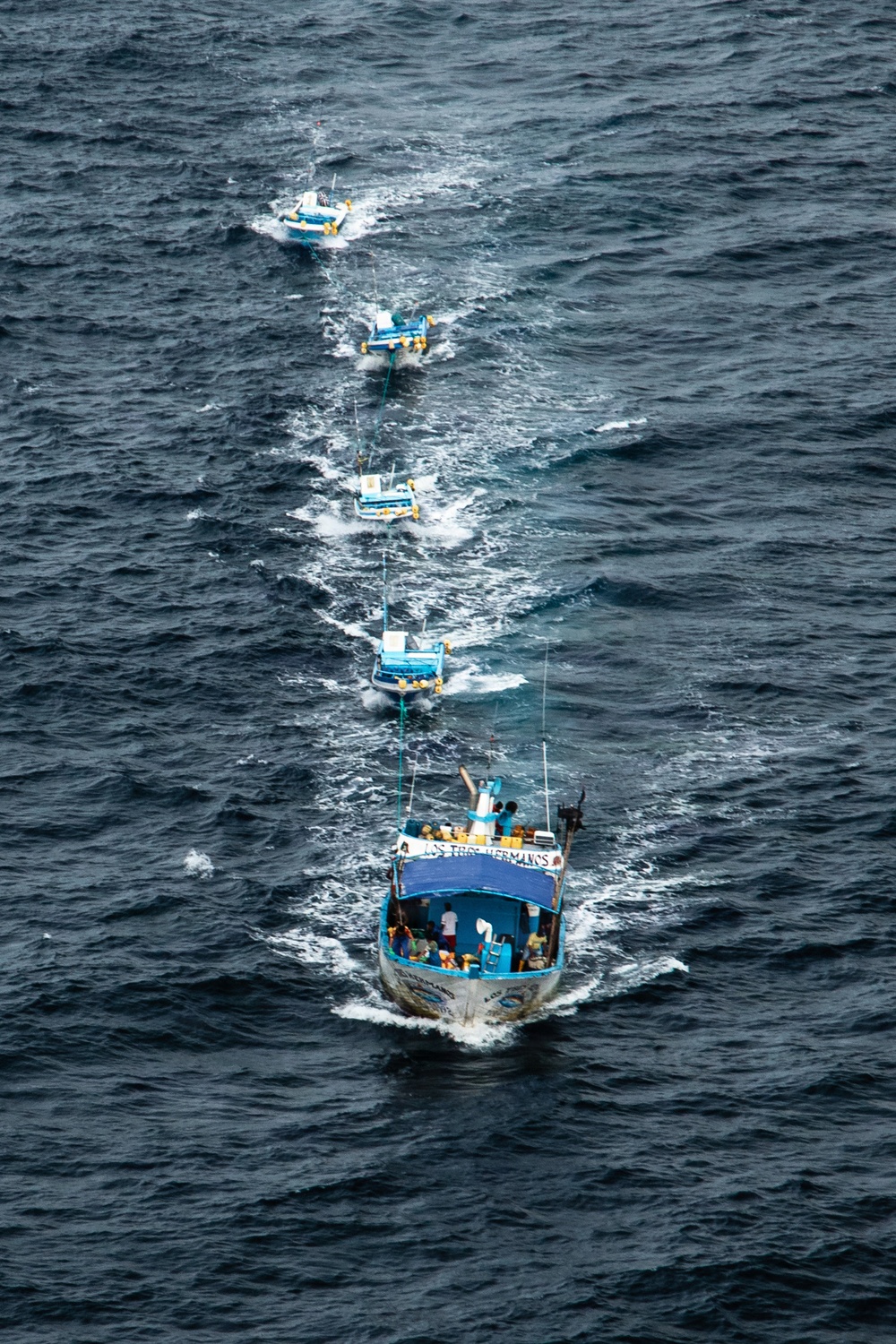 An aerial photograph shows a lead fishing vessel named 'Los Tres Hermanos' towing a chain of four smaller boats across open ocean. The boats are connected in a single-file line via tow ropes, all moving in the same direction through choppy dark blue-gray water. The image is taken from a significant elevation, likely from an aircraft or helicopter.