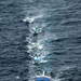 An aerial photograph shows a lead fishing vessel named 'Los Tres Hermanos' towing a chain of four smaller boats across open ocean. The boats are connected in a single-file line via tow ropes, all moving in the same direction through choppy dark blue-gray water. The image is taken from a significant elevation, likely from an aircraft or helicopter.