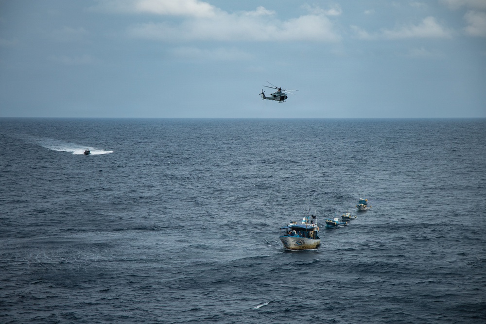 A maritime operation is underway on open ocean waters, showing a military or coast guard helicopter hovering above a convoy of several small fishing-style vessels moving in a line formation. A separate fast-moving patrol boat is visible in the background creating a white wake. The scene suggests an interdiction, escort, or law enforcement operation at sea.