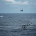A maritime operation is underway on open ocean waters, showing a military or coast guard helicopter hovering above a convoy of several small fishing-style vessels moving in a line formation. A separate fast-moving patrol boat is visible in the background creating a white wake. The scene suggests an interdiction, escort, or law enforcement operation at sea.