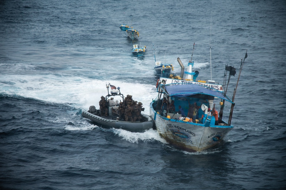 An aerial photograph shows a military or law enforcement rigid inflatable boat (RIB) intercepting a fishing vessel named 'Los Tres Hermanos' on open ocean waters. Armed personnel in tactical gear are aboard the RIB and appear to be conducting a maritime interdiction operation. Additional vessels are visible in the background, suggesting a coordinated multi-vessel operation.