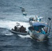 An aerial photograph shows a military or law enforcement rigid inflatable boat (RIB) intercepting a fishing vessel named 'Los Tres Hermanos' on open ocean waters. Armed personnel in tactical gear are aboard the RIB and appear to be conducting a maritime interdiction operation. Additional vessels are visible in the background, suggesting a coordinated multi-vessel operation.