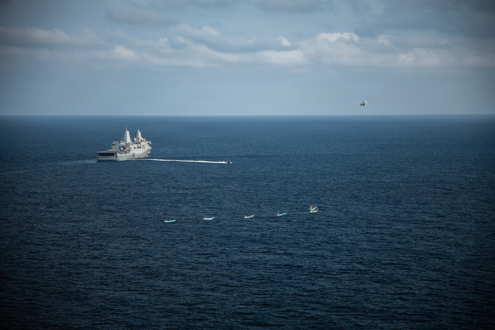 An aerial photograph shows a large naval warship, likely an amphibious transport dock vessel, underway at sea with a fast-moving smaller vessel creating a white wake nearby. Several small civilian or fishing boats are clustered in the foreground, while a helicopter is visible in the upper right portion of the sky. The scene suggests a maritime interdiction or patrol operation in open ocean.