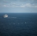 An aerial photograph shows a large naval warship, likely an amphibious transport dock vessel, underway at sea with a fast-moving smaller vessel creating a white wake nearby. Several small civilian or fishing boats are clustered in the foreground, while a helicopter is visible in the upper right portion of the sky. The scene suggests a maritime interdiction or patrol operation in open ocean.