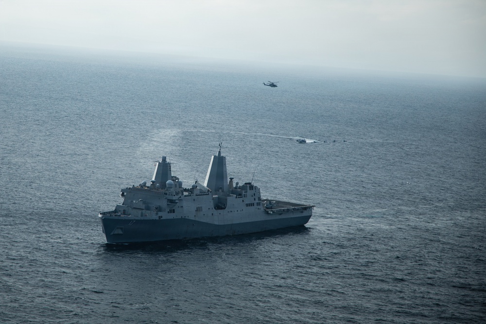 An aerial photograph shows a large gray naval warship, consistent with a San Antonio-class amphibious transport dock, underway on open ocean under overcast skies. A military helicopter is visible in the background, and what appears to be a smaller vessel or craft is operating nearby in the water. The scene suggests an active naval operation or exercise.