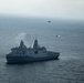 An aerial photograph shows a large gray naval warship, consistent with a San Antonio-class amphibious transport dock, underway on open ocean under overcast skies. A military helicopter is visible in the background, and what appears to be a smaller vessel or craft is operating nearby in the water. The scene suggests an active naval operation or exercise.