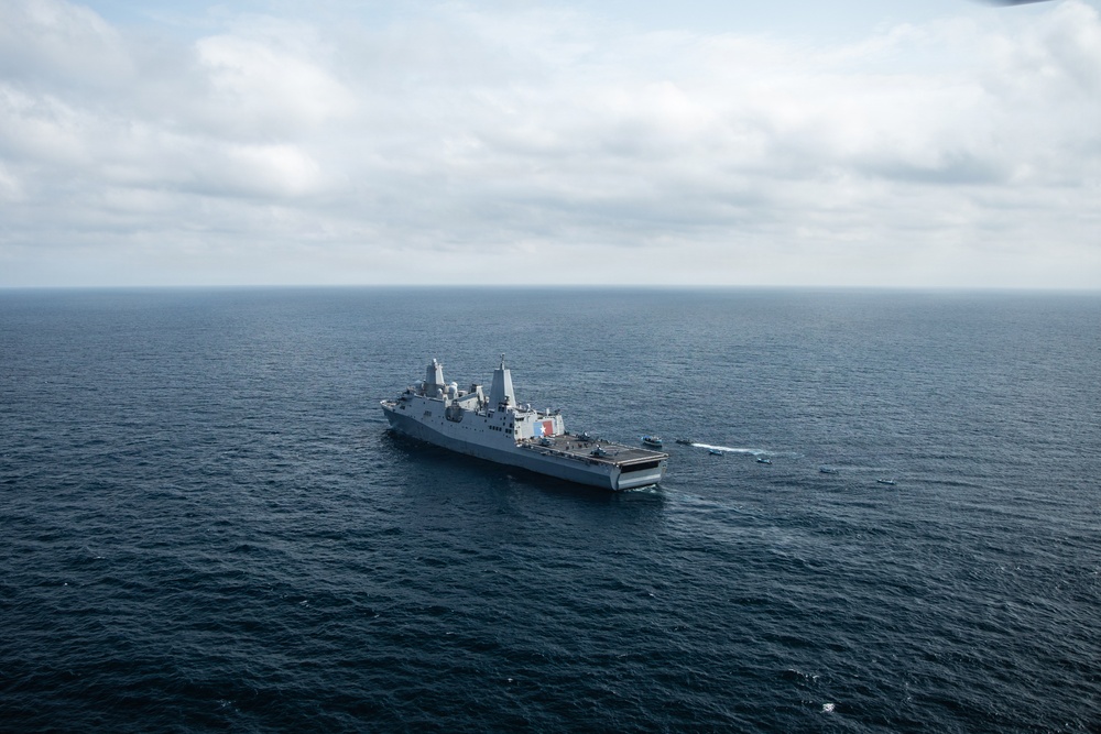 An aerial photograph of a large gray naval warship, consistent with a San Antonio-class amphibious transport dock (LPD), underway in open ocean. Several smaller vessels are visible alongside and near the stern of the ship, suggesting active small boat operations. The image was taken from an elevated vantage point, likely from a helicopter or aircraft.