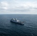 An aerial photograph of a large gray naval warship, consistent with a San Antonio-class amphibious transport dock (LPD), underway in open ocean. Several smaller vessels are visible alongside and near the stern of the ship, suggesting active small boat operations. The image was taken from an elevated vantage point, likely from a helicopter or aircraft.