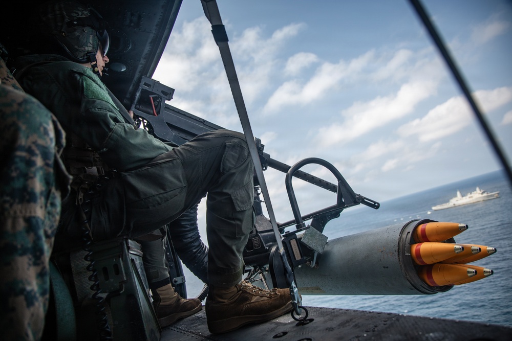 A military aircrew member, wearing a flight helmet and camouflage uniform, is seated in the open door of a military helicopter over open ocean. Visible in the foreground is a rocket pod loaded with what appear to be practice or training rockets with orange tips. A naval vessel is visible in the background on the water.