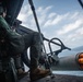 A military aircrew member, wearing a flight helmet and camouflage uniform, is seated in the open door of a military helicopter over open ocean. Visible in the foreground is a rocket pod loaded with what appear to be practice or training rockets with orange tips. A naval vessel is visible in the background on the water.