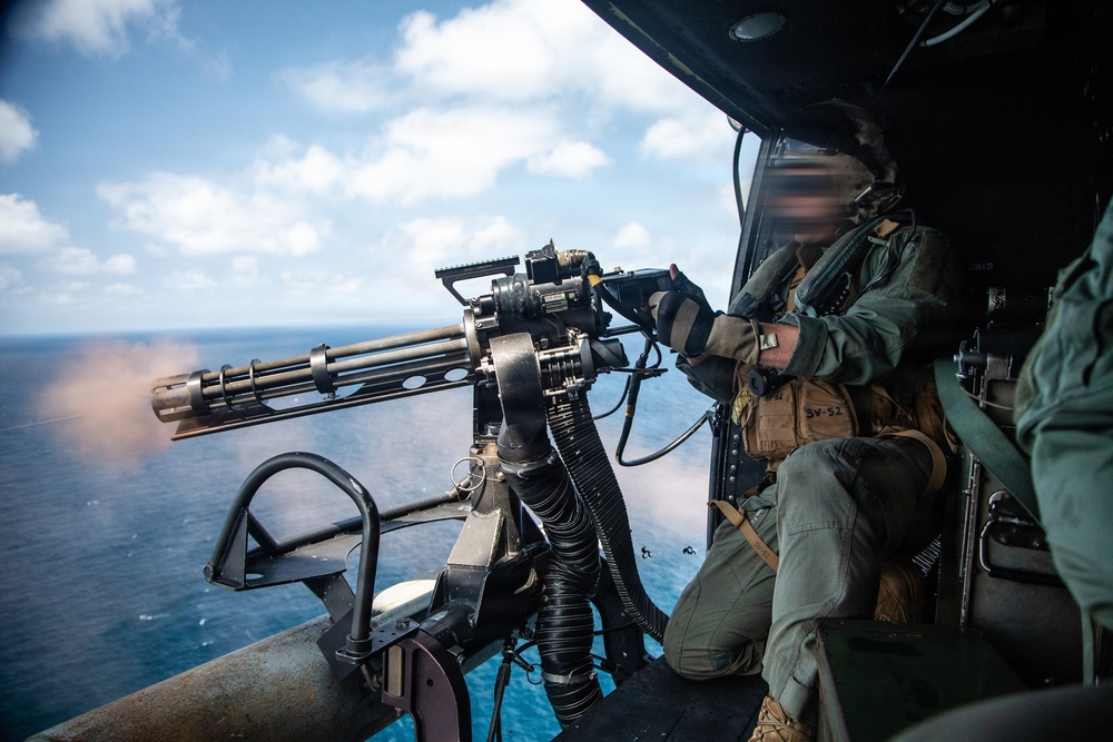 A military aircrew member is operating a multi-barrel rotary machine gun (consistent with an M134 Minigun) from the open door of a helicopter over an ocean environment. The weapon appears to be actively firing, as evidenced by visible muzzle flash and smoke at the barrel end. The operator is wearing flight gear including a helmet with visor and a survival vest marked 'SV-52'.