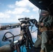 A military aircrew member is operating a multi-barrel rotary machine gun (consistent with an M134 Minigun) from the open door of a helicopter over an ocean environment. The weapon appears to be actively firing, as evidenced by visible muzzle flash and smoke at the barrel end. The operator is wearing flight gear including a helmet with visor and a survival vest marked 'SV-52'.