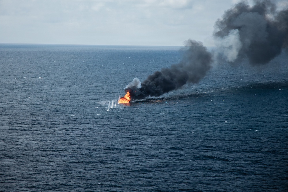 An open-water fire is burning on the ocean surface, producing a large column of dense black smoke that drifts to the upper right of the frame. The fire appears to be a controlled or in-situ burn, possibly of an oil spill or a vessel, based on the dark, hydrocarbon-characteristic smoke. The image is taken from an elevated aerial perspective over open, deep-blue ocean water.