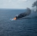 An open-water fire is burning on the ocean surface, producing a large column of dense black smoke that drifts to the upper right of the frame. The fire appears to be a controlled or in-situ burn, possibly of an oil spill or a vessel, based on the dark, hydrocarbon-characteristic smoke. The image is taken from an elevated aerial perspective over open, deep-blue ocean water.