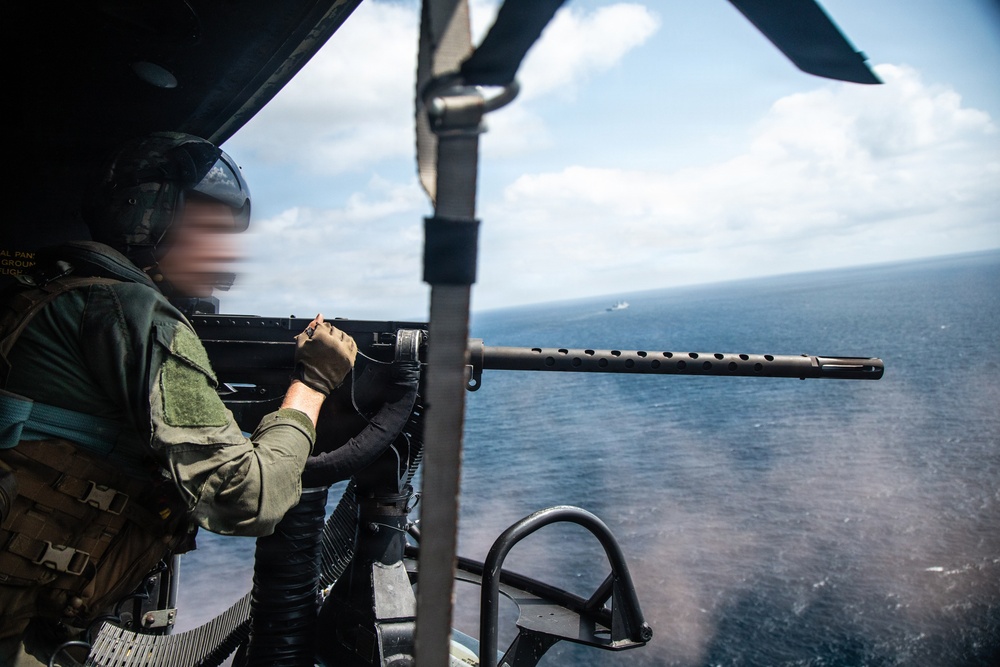 A military aircrew member is operating a door-mounted machine gun from what appears to be a helicopter over open ocean. The gunner is wearing flight gear including a helmet and tactical vest, and is positioned at an open door with the weapon aimed outward. A vessel is faintly visible on the horizon in the background.