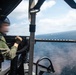 A military aircrew member is operating a door-mounted machine gun from what appears to be a helicopter over open ocean. The gunner is wearing flight gear including a helmet and tactical vest, and is positioned at an open door with the weapon aimed outward. A vessel is faintly visible on the horizon in the background.