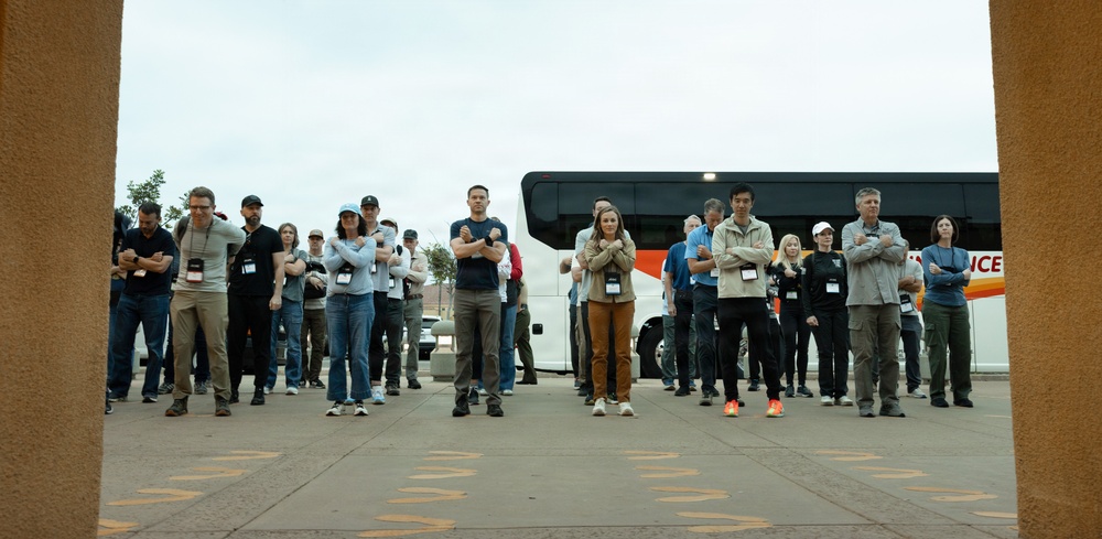 A large group of approximately 20-30 people stand in a row outdoors, most with their arms crossed, facing the camera. They appear to be participants in an organized event or training, many wearing lanyards with badges. A white coach bus is visible in the background, and the scene is framed by what appears to be an adobe or stucco architectural element in the foreground.