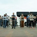 A large group of approximately 20-30 people stand in a row outdoors, most with their arms crossed, facing the camera. They appear to be participants in an organized event or training, many wearing lanyards with badges. A white coach bus is visible in the background, and the scene is framed by what appears to be an adobe or stucco architectural element in the foreground.
