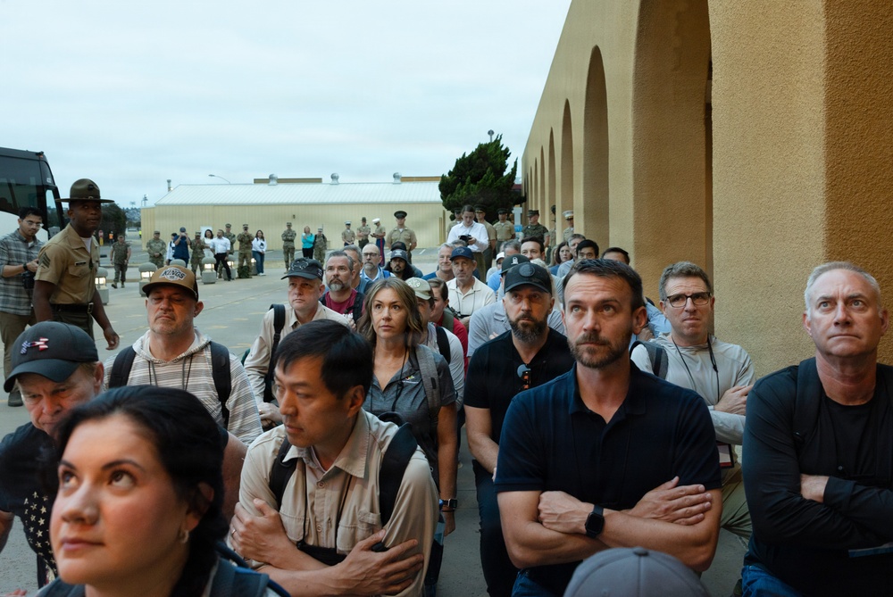 A large group of civilians and military personnel are gathered outdoors near a tan/beige building with arched architectural details, likely on a military base. The crowd appears to be attentively watching or listening to something off-camera, with many individuals standing with arms crossed. The setting and mix of uniformed and civilian attendees suggests an official event, demonstration, or briefing.
